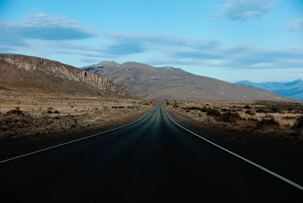 A black asphalt road surrounded by green and brown mountains in El Calafate, Argentina and blue sky with white clouds.