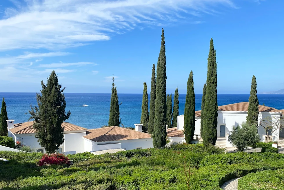 Green grass, long trimmed trees, white houses with blue sea in the background.