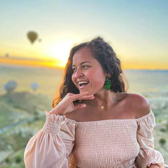 Travel Advisor Rosealety Galea stands on a balcony at sunset wearing a light pink dress with hot air balloons floating through the sky. 