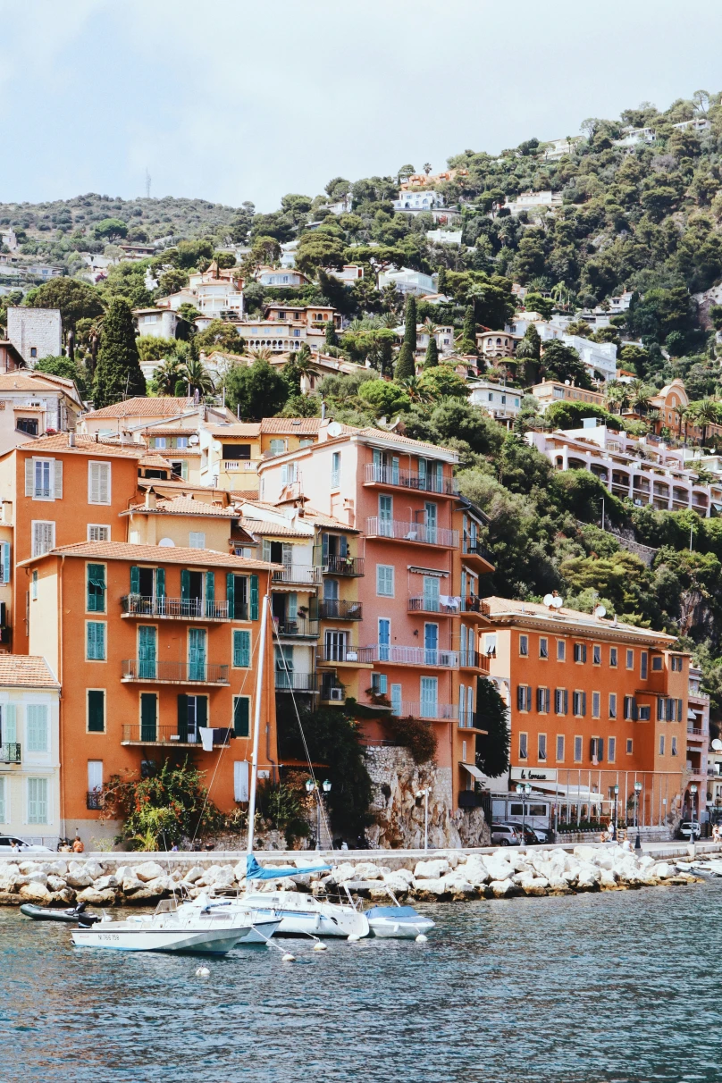 A view of the coastline with orange buildings and lush green landscape on the hillside with a boat sailing in view