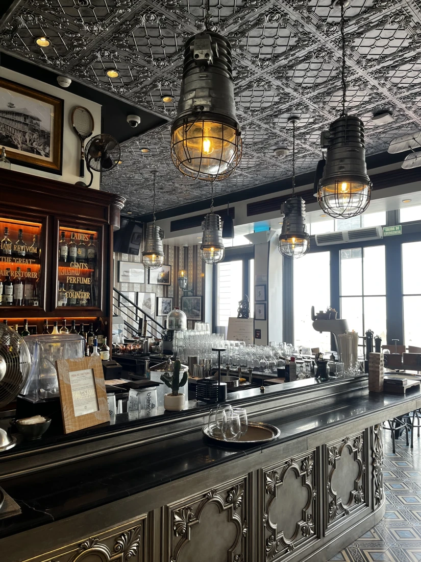 A restaurant bar with hanging lights, detailed ceiling and large windows in the back right corner near a staircase. 