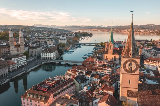 An overhead view of Zurich in the daytime with a canal, bridges, old stone buildings, church towers and mountain range in the distance. 