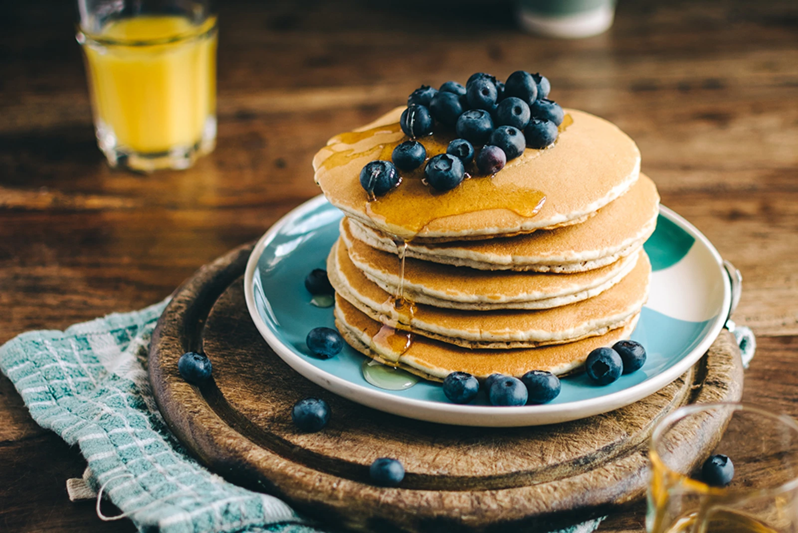stack of five golden pancakes with blueberries and maple syrup on a blue plate and wooden platter orange juice in clear glass