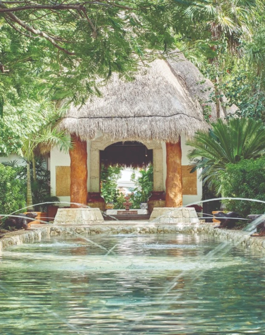 a turquoise reflection pool leading up to a thatched-roof structure in the jungle