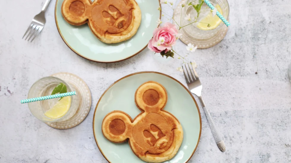 Mickey Mouse shaped pancakes in tea plates with water and utensils on the side.