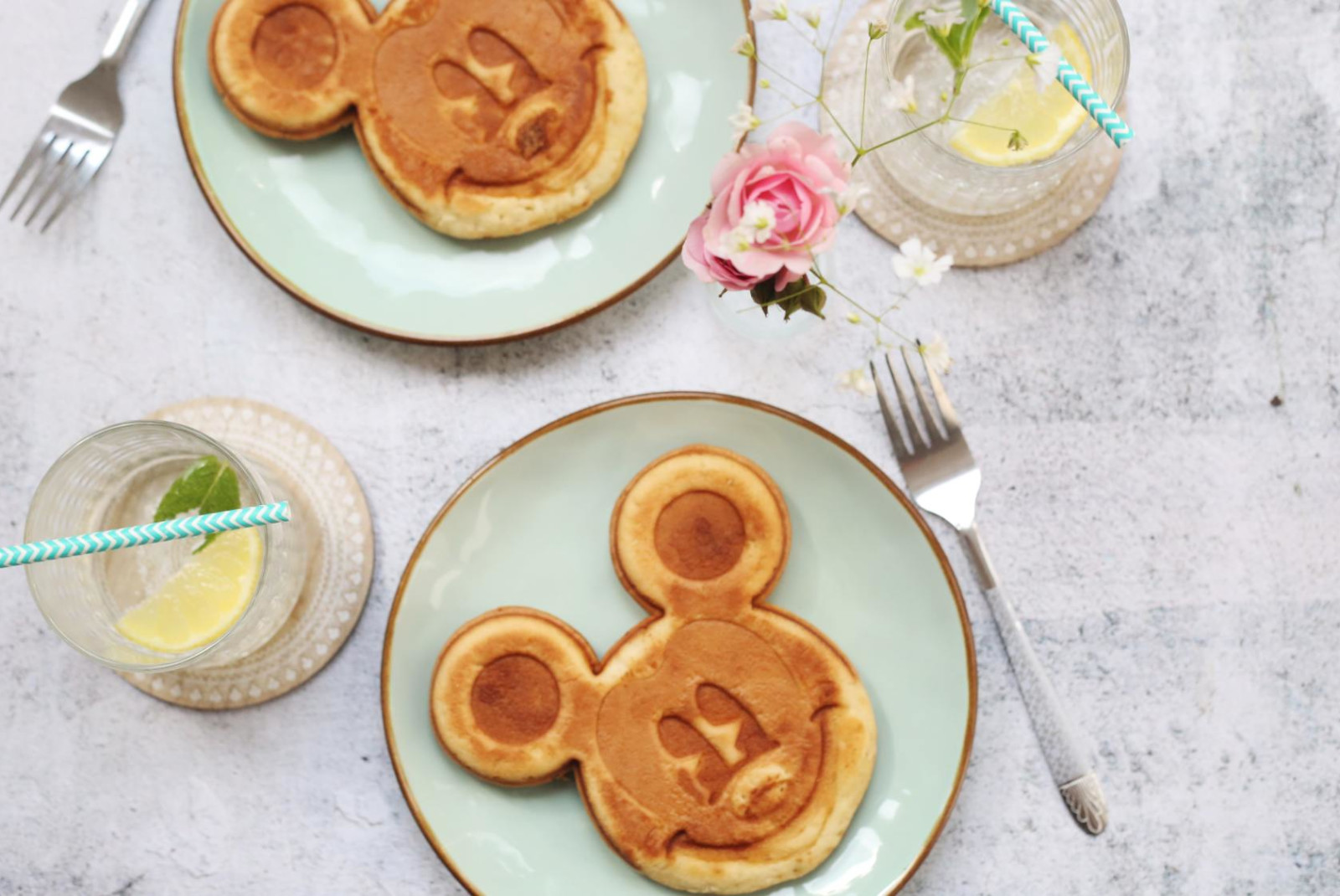 Mickey Mouse shaped pancakes in tea plates with water and utensils on the side.