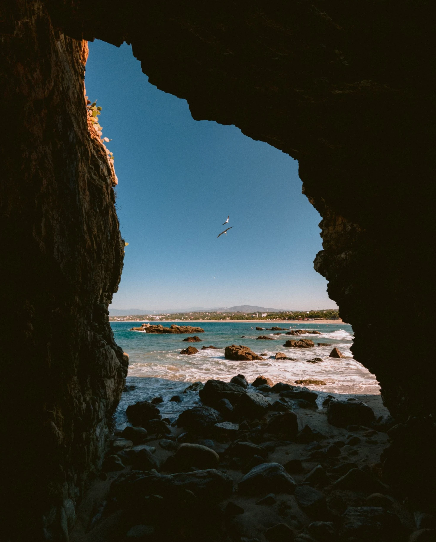 view through a rock formation of a rocky beach on a blue day with a hang glider 