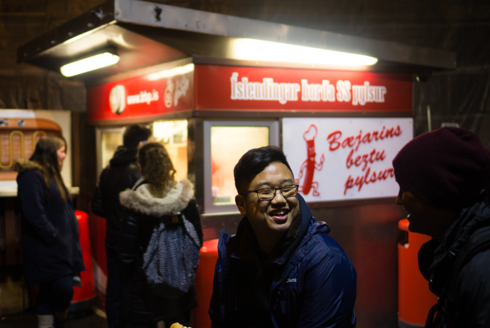 A red and white famous hot dog stand in this Iceland Itinerary with five people in dark coats waiting in line.