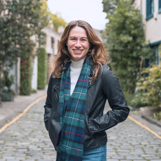 Fora travel agent Misha Sharp wearing jeans and plaid scarf with buildings and cobblestone street in background during daytime