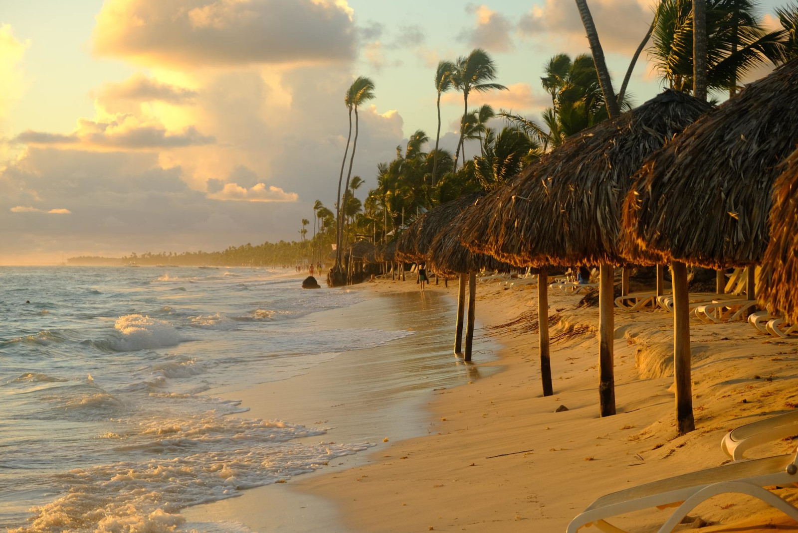 Beach in Punta Cana during sunset, reflecting light against clouds, ocean and sand with straw huts and palm trees.