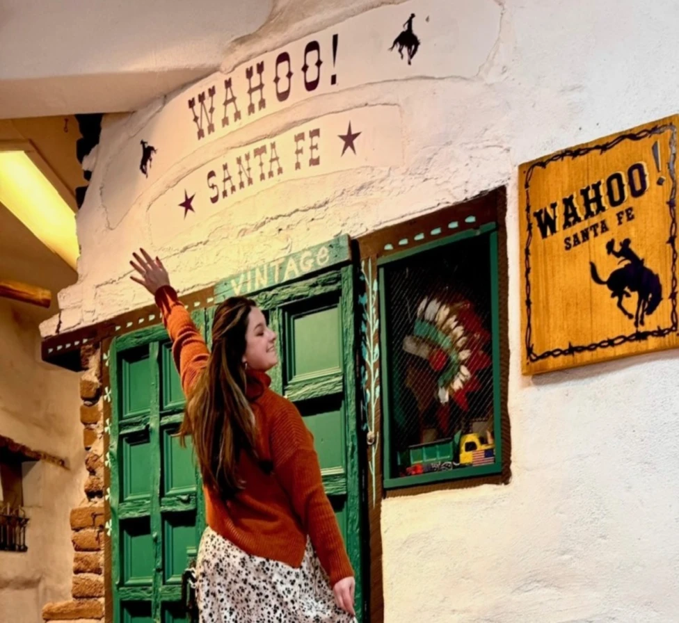 Woman posing in front of a white all with different frames and wall art saying 'Wahoo! Santa Fe'