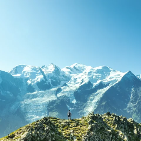 A man on a green mountain top looking out toward snow covered peaks. 