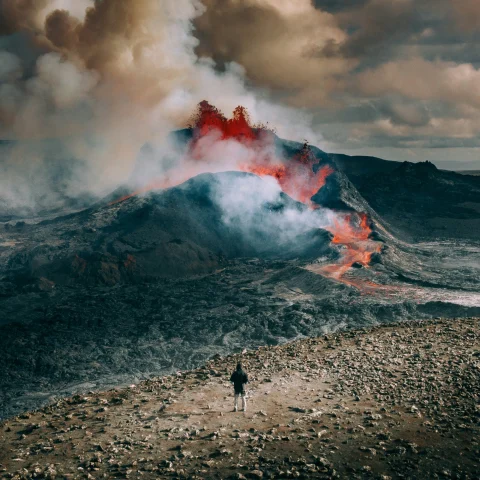 A person standing on rocky terrain with an active volcano and eruptions of smoke in the background. 