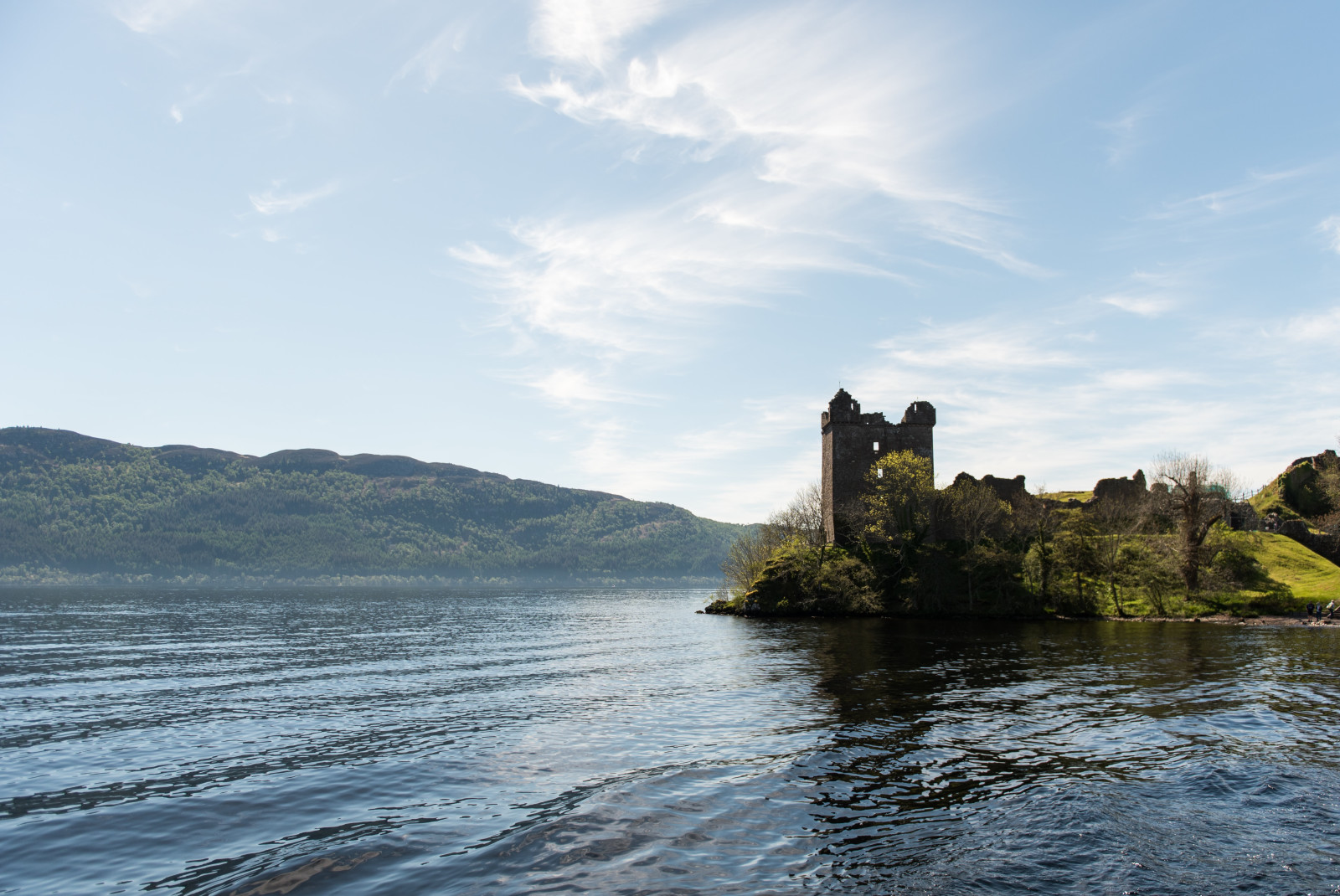 Loch Ness lake in Scotland with blue calm waters and brown castle ruins and on a hill with green grass and more hills in the distance.