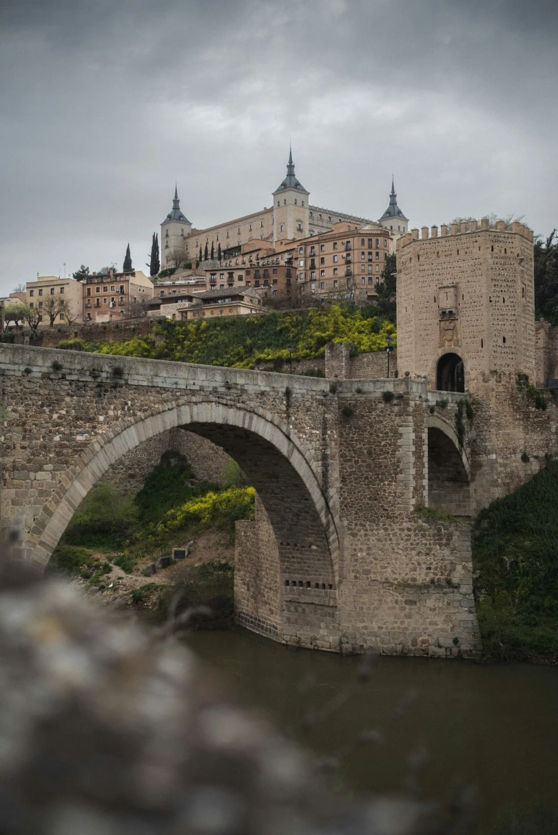 An old-world, stone castle perched on a hill, with a stone bridge over a narrow river in front of it on a cloudy day. 