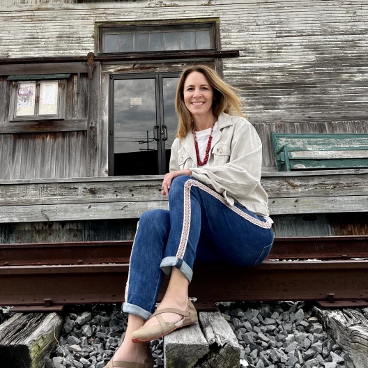 Fora travel agent Laura Cox wearing blue pants and sandals sits on railroad track with wooden building in background