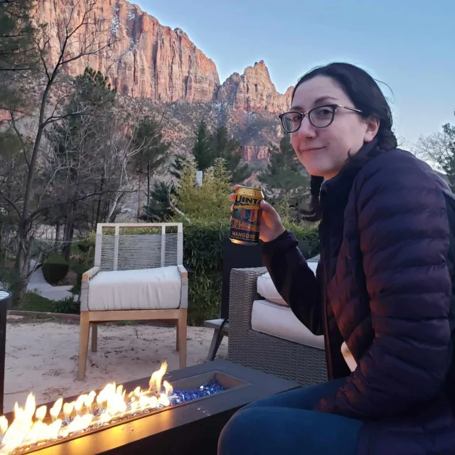 Picture of Mary in a black puffer jacket, having a drink in front of a fire pit with red mountains in the background.