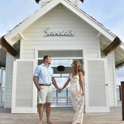 Couple in beach attire in front of a white Sandals building with a view of the ocean in the background