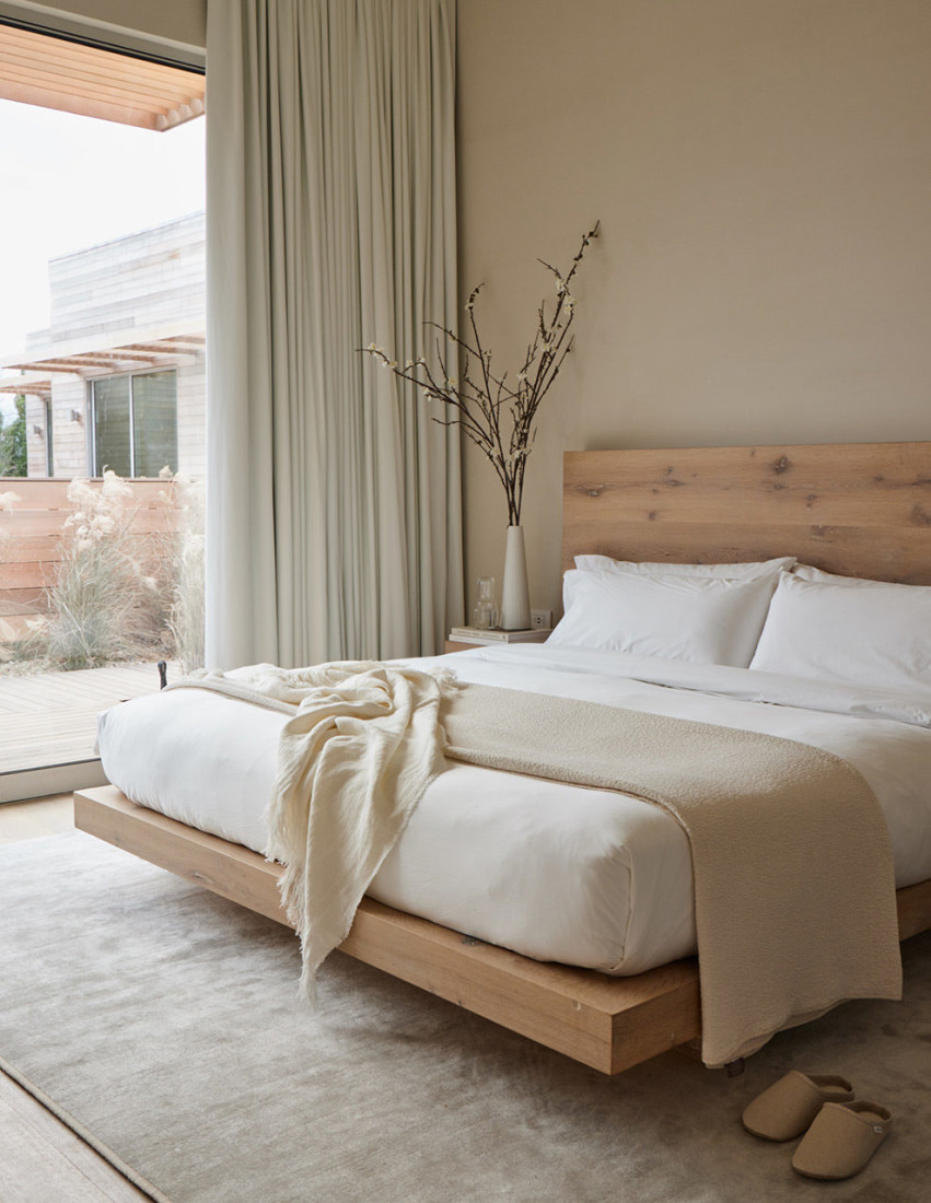 a luxe white bed with a wooden headboard in a room with floor-to-ceiling windows