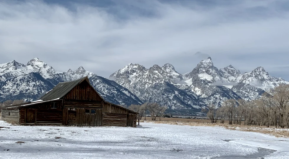 Wood barn with snowy yard with mountains in the background during daytime