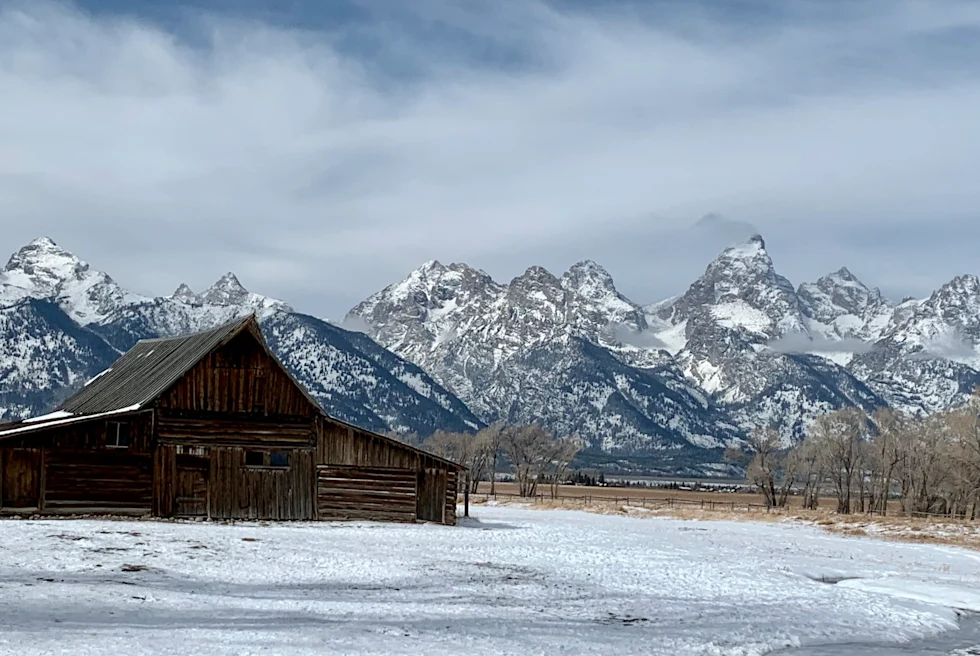 Wood barn with snowy yard with mountains in the background during daytime