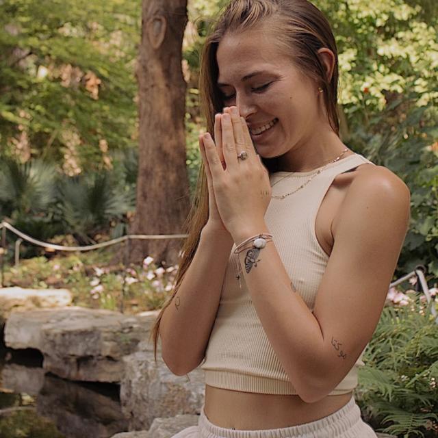 Fora travel agent Taylor Medows wears tan tank top and sits in prayer with trees in the background