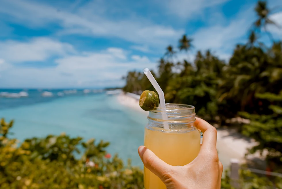 a hand holds a cocktail over a sandy sunny beach view