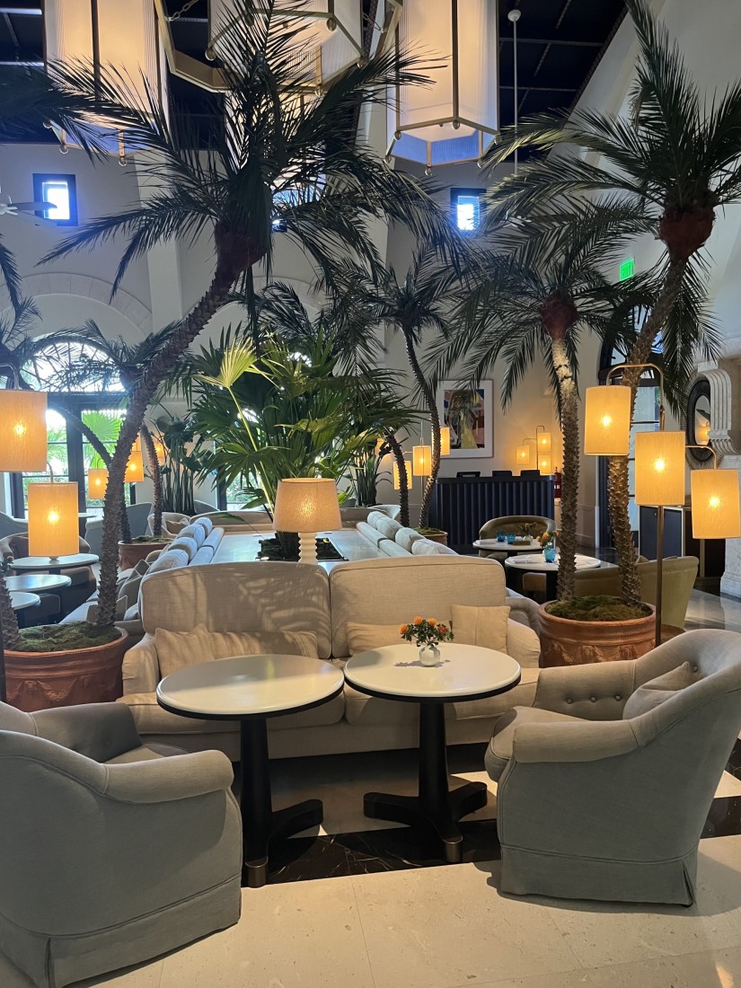 A hotel lobby with tall palm trees, lounge seating, round coffee tables and various lanterns turned on beneath large hanging chandeliers. 
