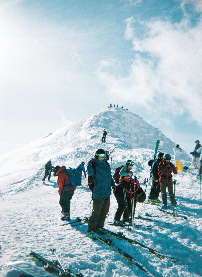 A group of people in ski gear standing on top of snowy ground with a snowy peak in the background while the sun shines down onto them. 