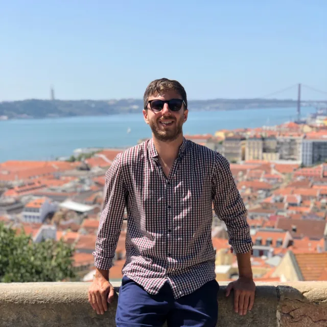 Travel Advisor Josh Volpe in a plaid shirt sitting on a wall overlooking Lisbon, Portugal.