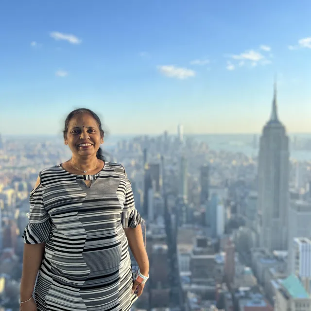 travel advisor Kalpana Shah stands at the top of a skyscraper overlooking New York City wearing jeans and a black and white striped t-shirt
