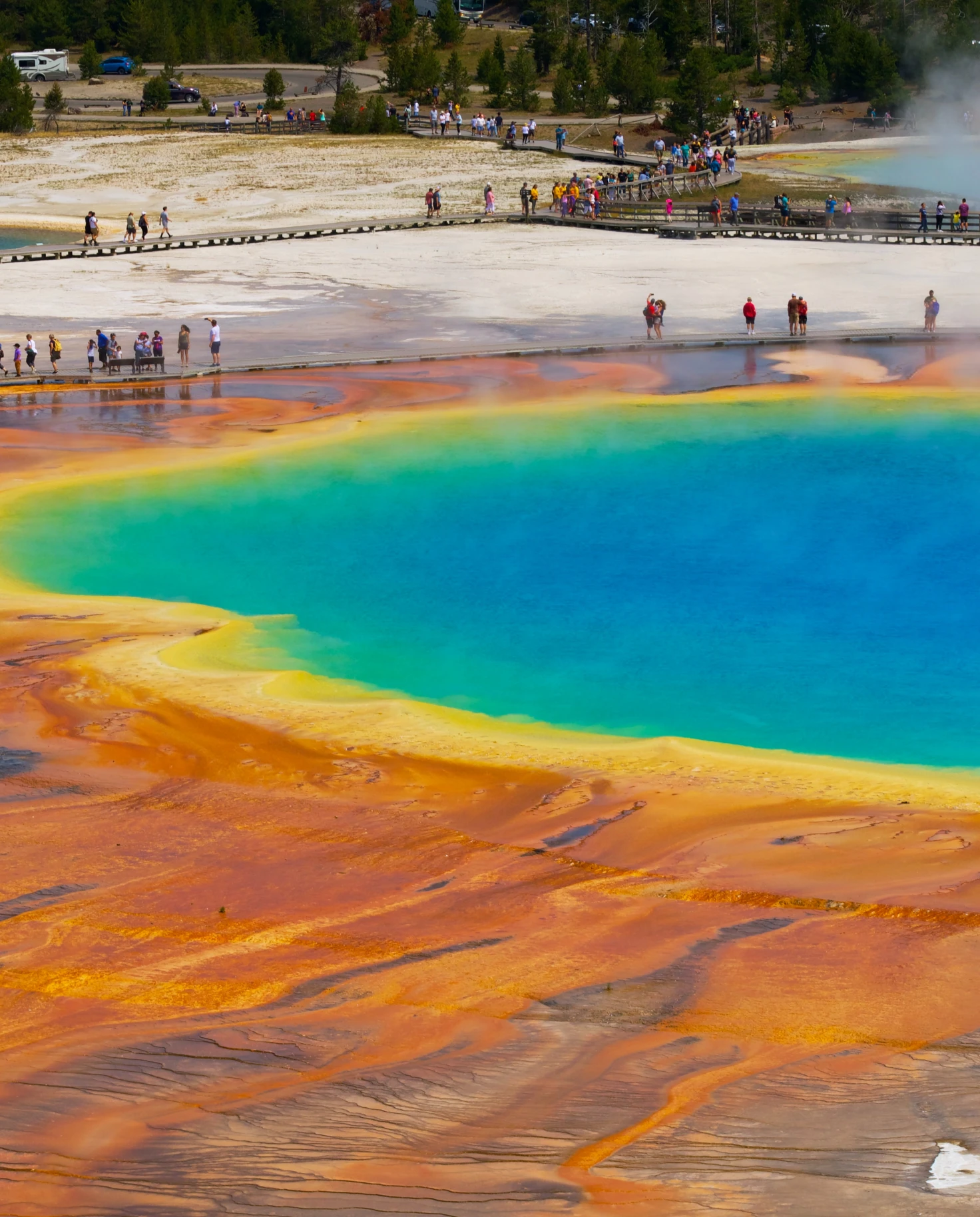 An orange, yellow, green and blue geysir in Yellowstone National Park with people standing around the outskirts.