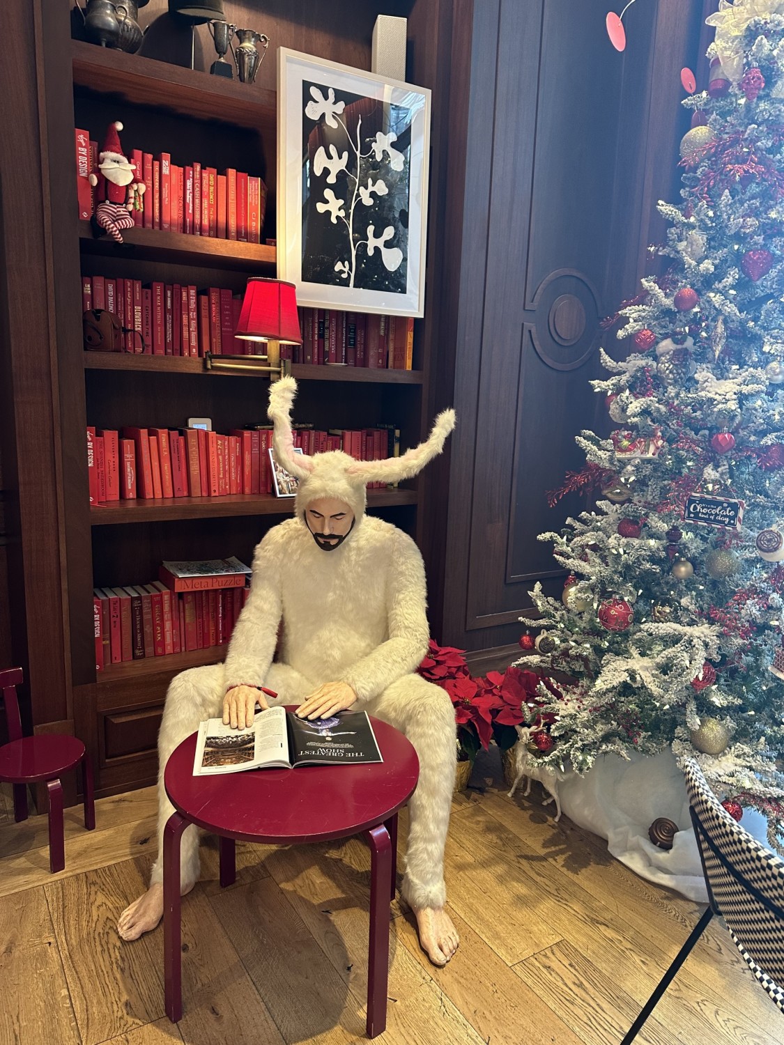 Unique artwork at the hotel of a man dressed in a furry bunny outfit sitting at a small red table reading. There is a bookshelf of red books behind and a christmas tree in view.