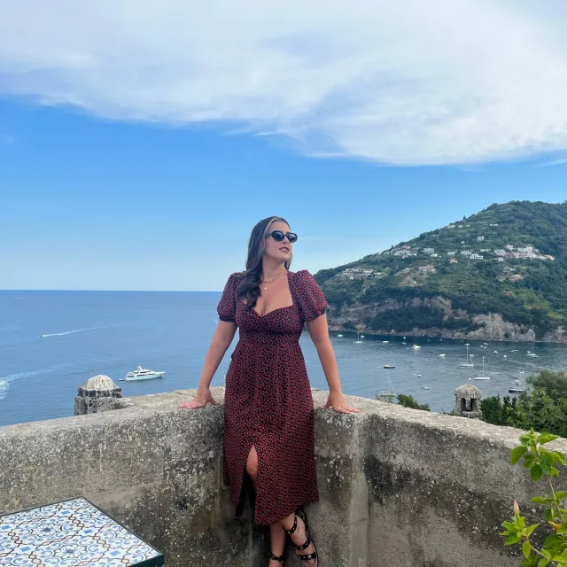 Travel Advisor Carissa Bartoli with a dark red dress in front of a ledge with water and green lush hills in the background.