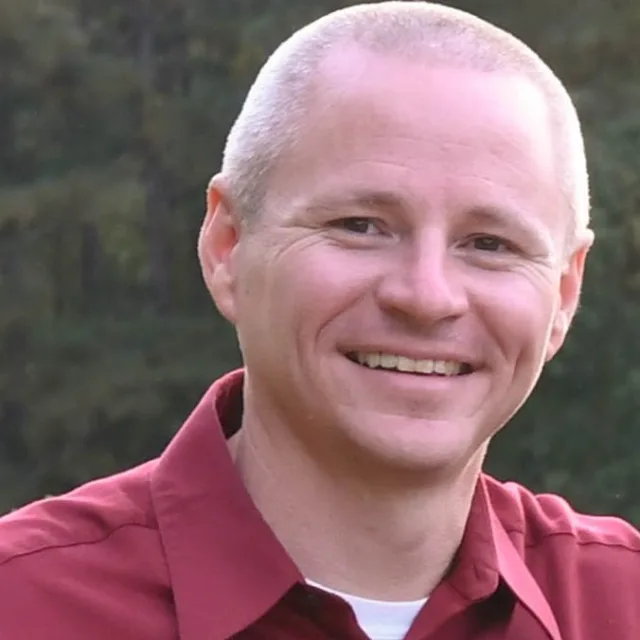 man with a white buzz cut and red collared shirt smiling