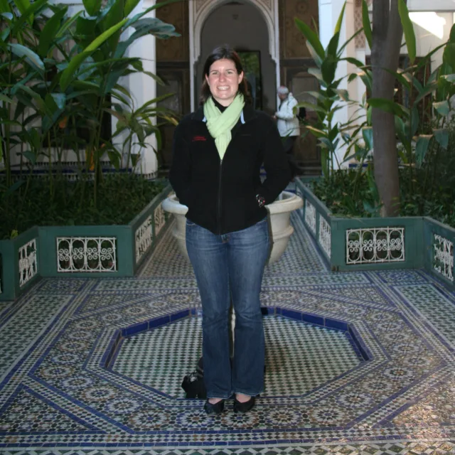 Picture of Lindsay wearing a black jacket, green scarf and jeans standing on a mosaic tile surrounded by palm leaf greenery