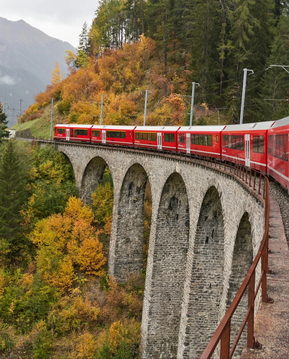 An image depicting a red train on a tall, stone railroad with autumnal trees in the surrounding area. 