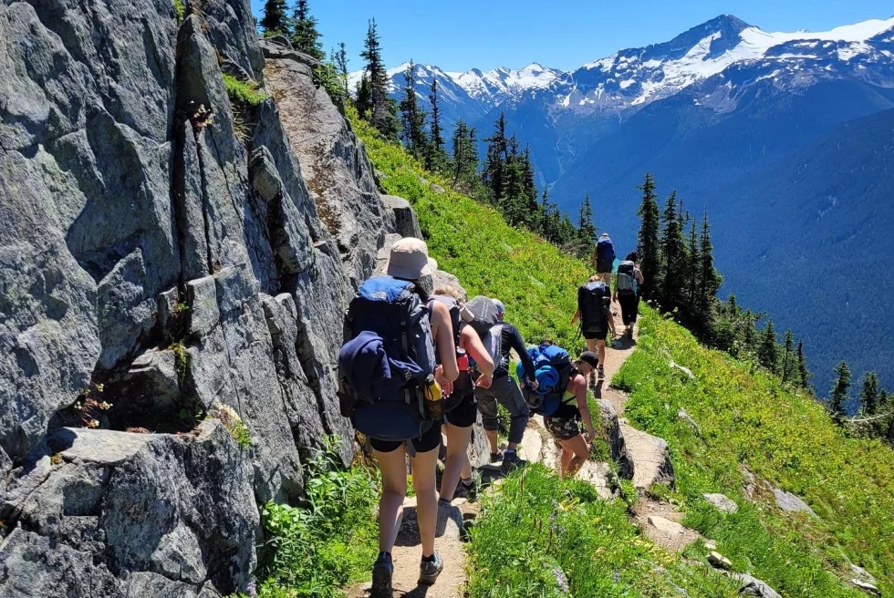 a group walks on a path on the side of a steep mountain with green foliage and snowy mountain range 