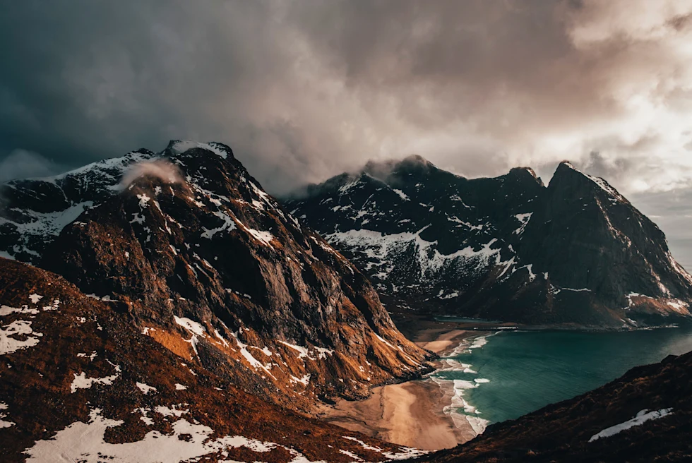 snowy mountains near body of water with cloudy skies