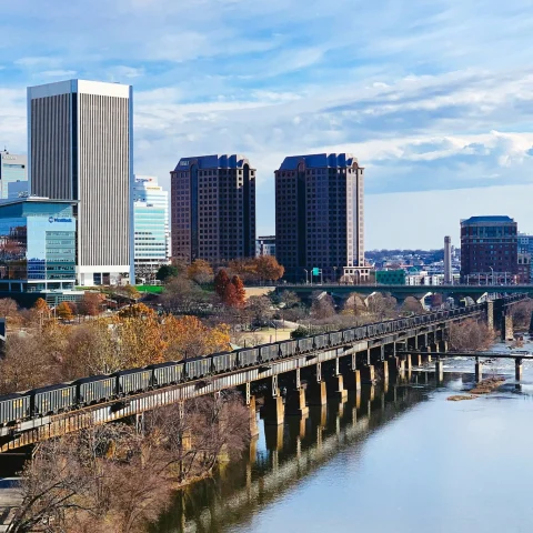 A view of Richmond, featuring a river, bridge, Autumnal trees and skyscrapers under a cloudy blue sky. 
