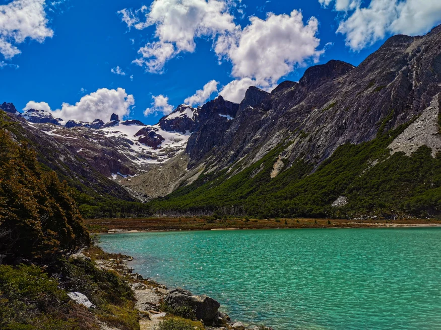 A blue-green glacial lake called Laguna Esmeralda in Argentina with tall dark grey rocky mountains with green trees lining them and blue sky with white clouds.