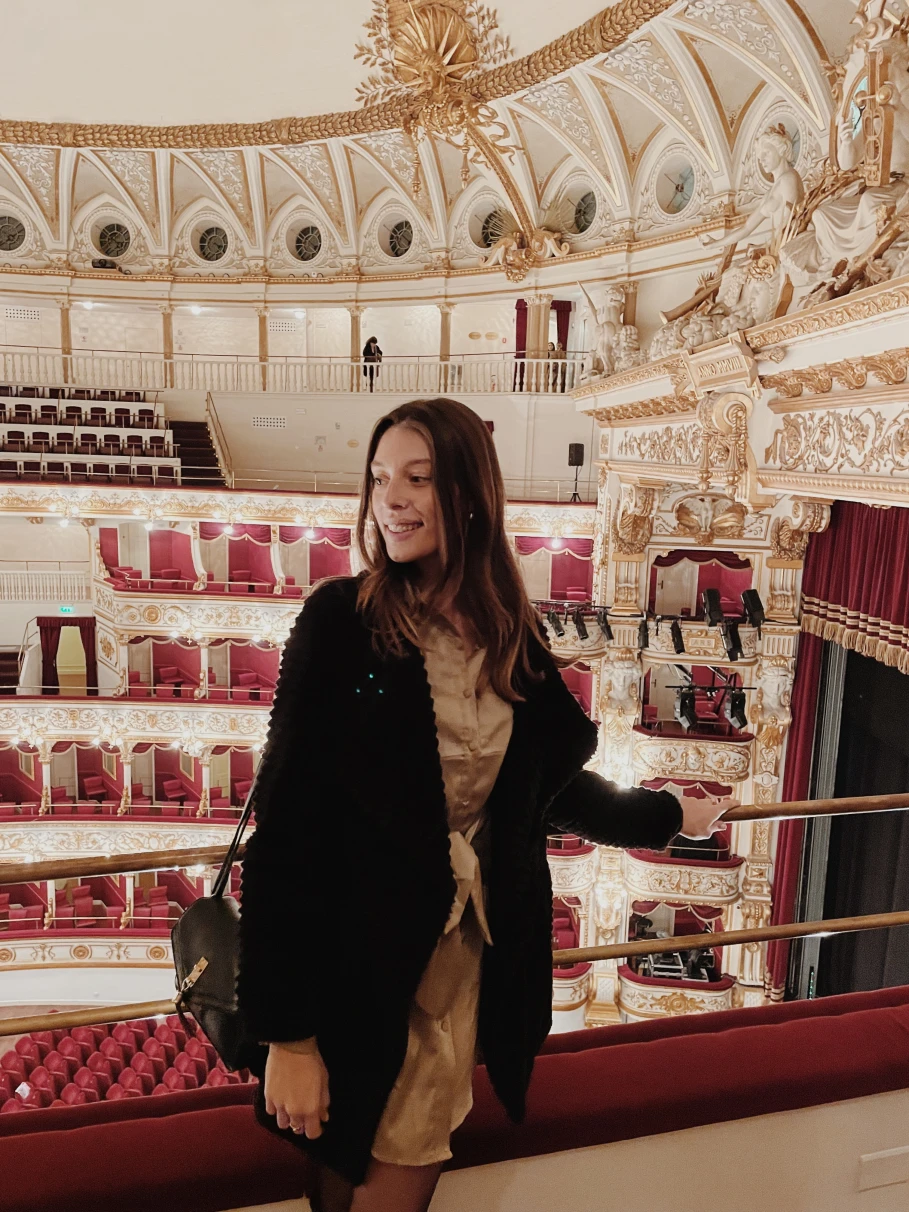 A girl posing for a photo inside of a glamorous old theatre with red seats and white and gold details. 