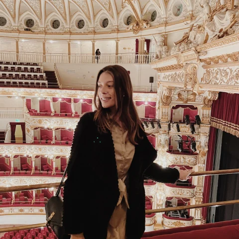 A girl posing for a photo inside of a glamorous old theatre with red seats and white and gold details. 