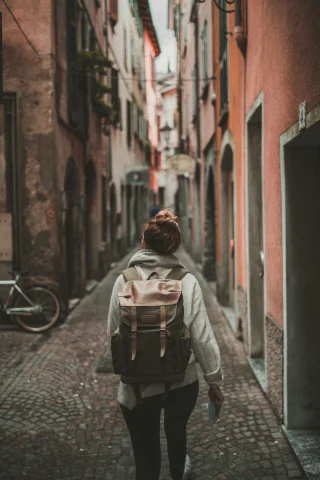 A person wearing a backpack walking down a narrow, cobblestone alley surrounded by colorful, European buildings. 