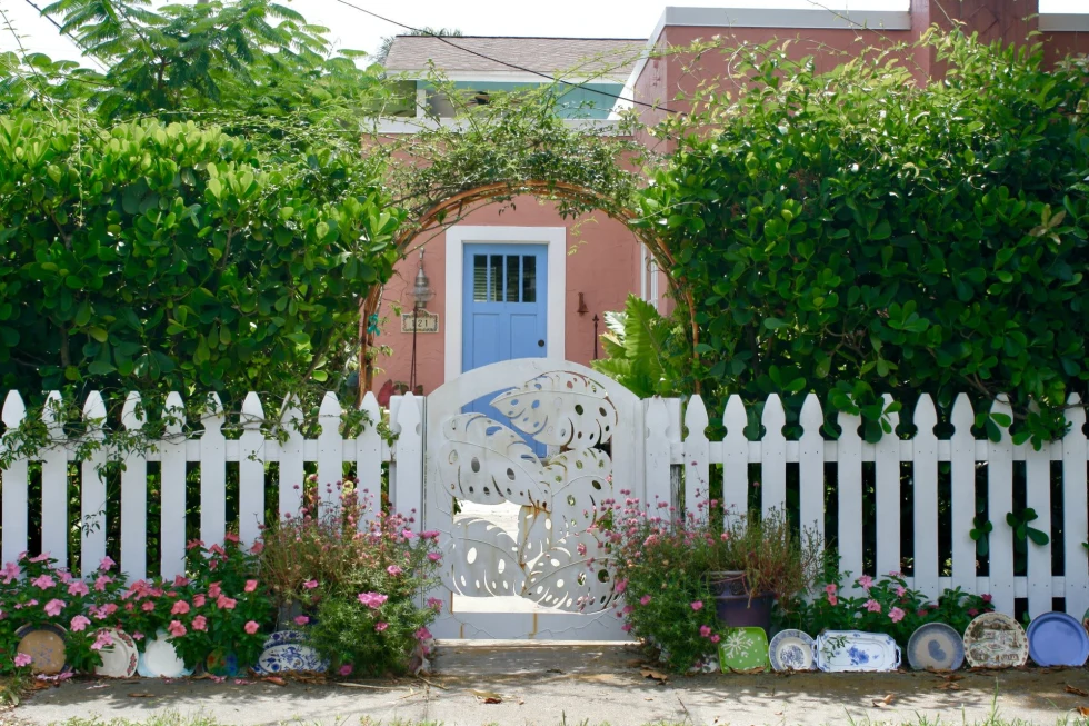a charming pink beach cottage with a blue door and white picket fence and arched bushes 
