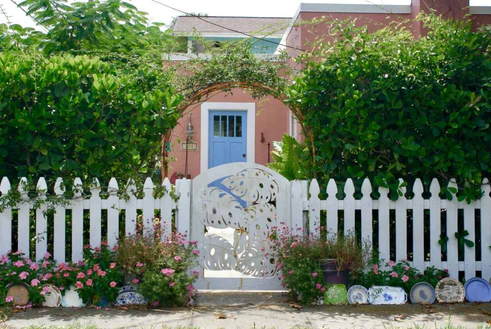 a charming pink beach cottage with a blue door and white picket fence and arched bushes 