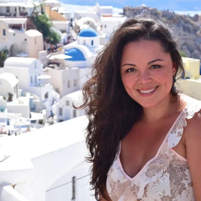Travel advisor Jamielys Ponce de Leon in a floral top standing in front of the white hillside buildings of Greece.