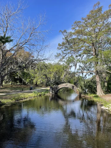 An image of a creek with a stone arch bridge in the distance, surrounded by tall trees with green leaves and green grass beneath a blue sky. 
