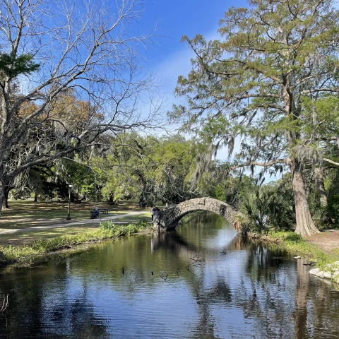 An image of a creek with a stone arch bridge in the distance, surrounded by tall trees with green leaves and green grass beneath a blue sky. 