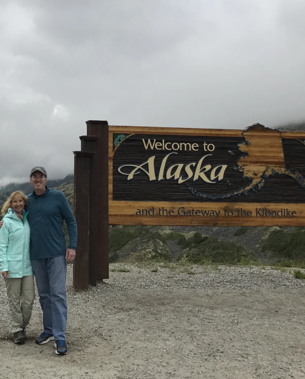 A couple standing in front of a "Welcome to Alaska" sign in front of a mountain landscape, during the best time to travel to Alaska.